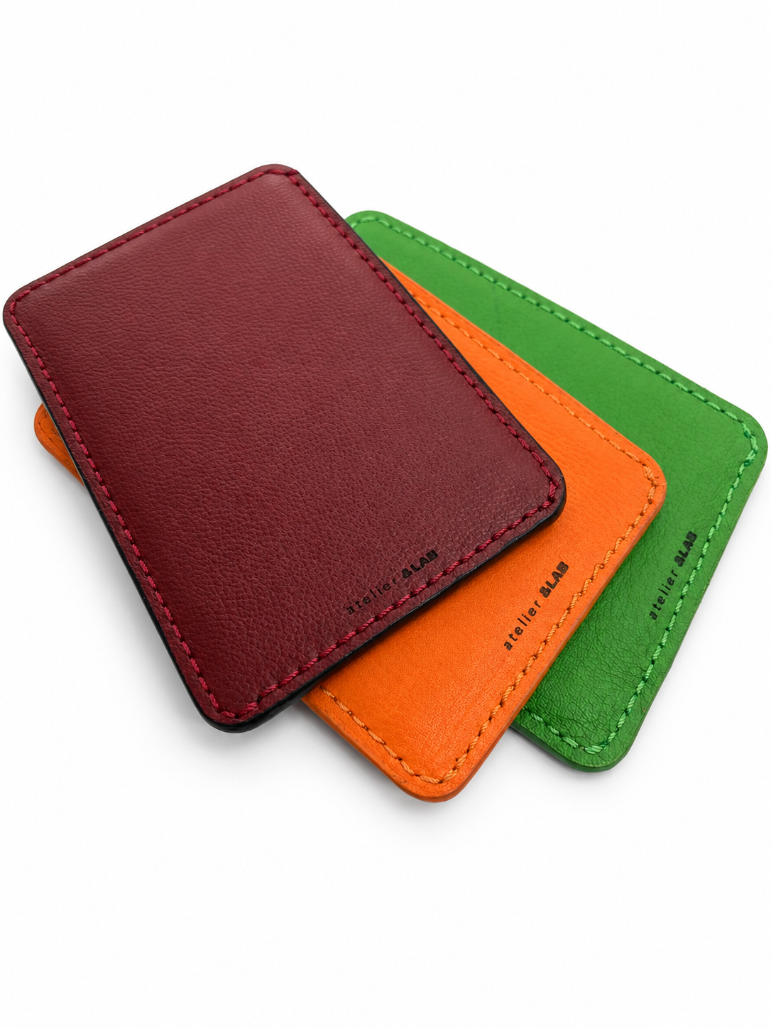 Back view of three overlapping MODULE Simple card holders by atelier &LAB in Bordeaux, orange and green upcycled full-grain calf leather, showing the leather texture, stitching and embossed brand logos. Designed and made near Paris.
